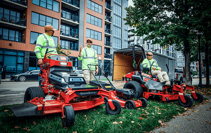 Landscape contractors with Toro GrandStand stand-on mower, walk-behind, and zero-turn equipment beside an equipment trailer near a commercial building